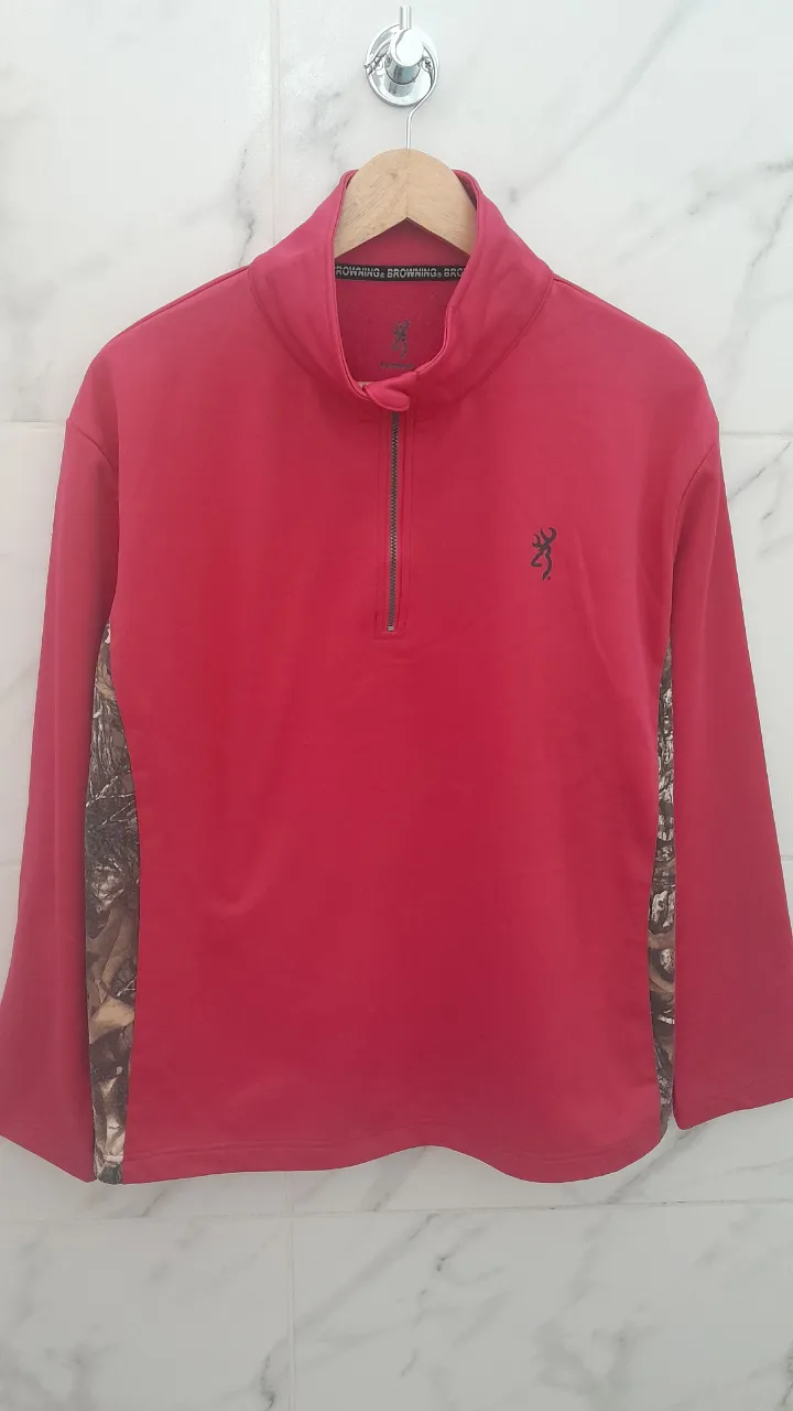 Buzo deportivo de color rosa con cuello alto y cierre parcial. Posee detalles de estampa camuflada en los laterales de las mangas. Logo de la marca bordado en el frente.