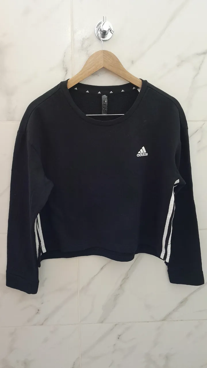Buzo crop negro con logo de Adidas blanco en el frente y tres tiras blancas en las mangas.