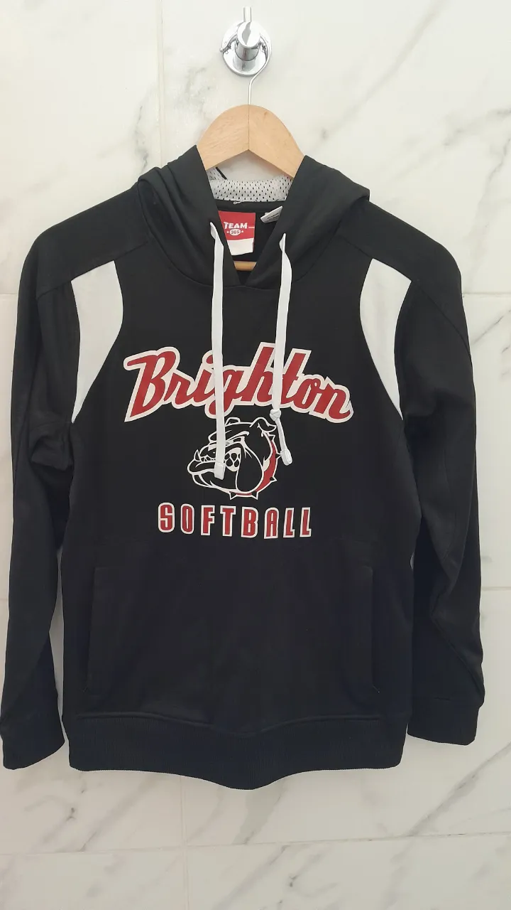 Buzo negro con capucha y cordones blancos. Estampado frontal con el texto "Brighton" y un logo de perro bulldog con la palabra "SOFTBALL". Tiene bolsillos con cierre a los costados. En la espalda tiene el número "6" estampado.