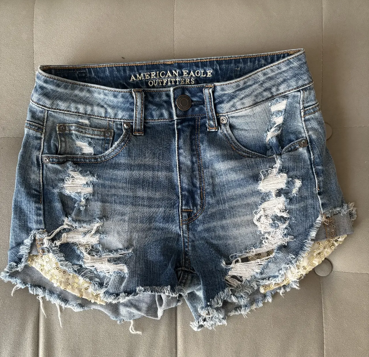 American Eagle Outfitters - Short de jean tiro alto, con detalles rotos y bordes deshilachados. Bolsillos con lentejuelas doradas. Ideal para un look casual y canchero.