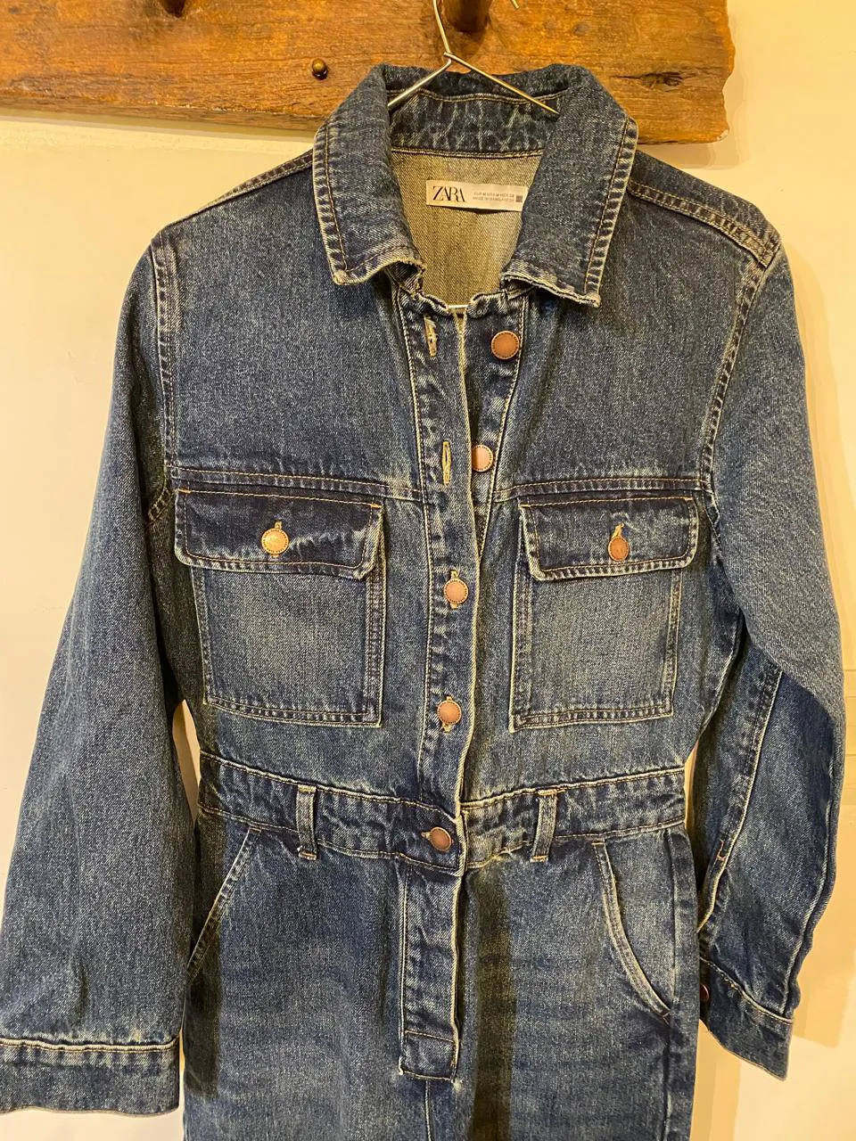 Mono de jean azul con cuello de camisa, mangas largas y botones al frente. Posee dos bolsillos superiores con tapa y botones, y dos bolsillos inferiores tipo ojal. Tiene un cinturón de tela de jean con hebilla.