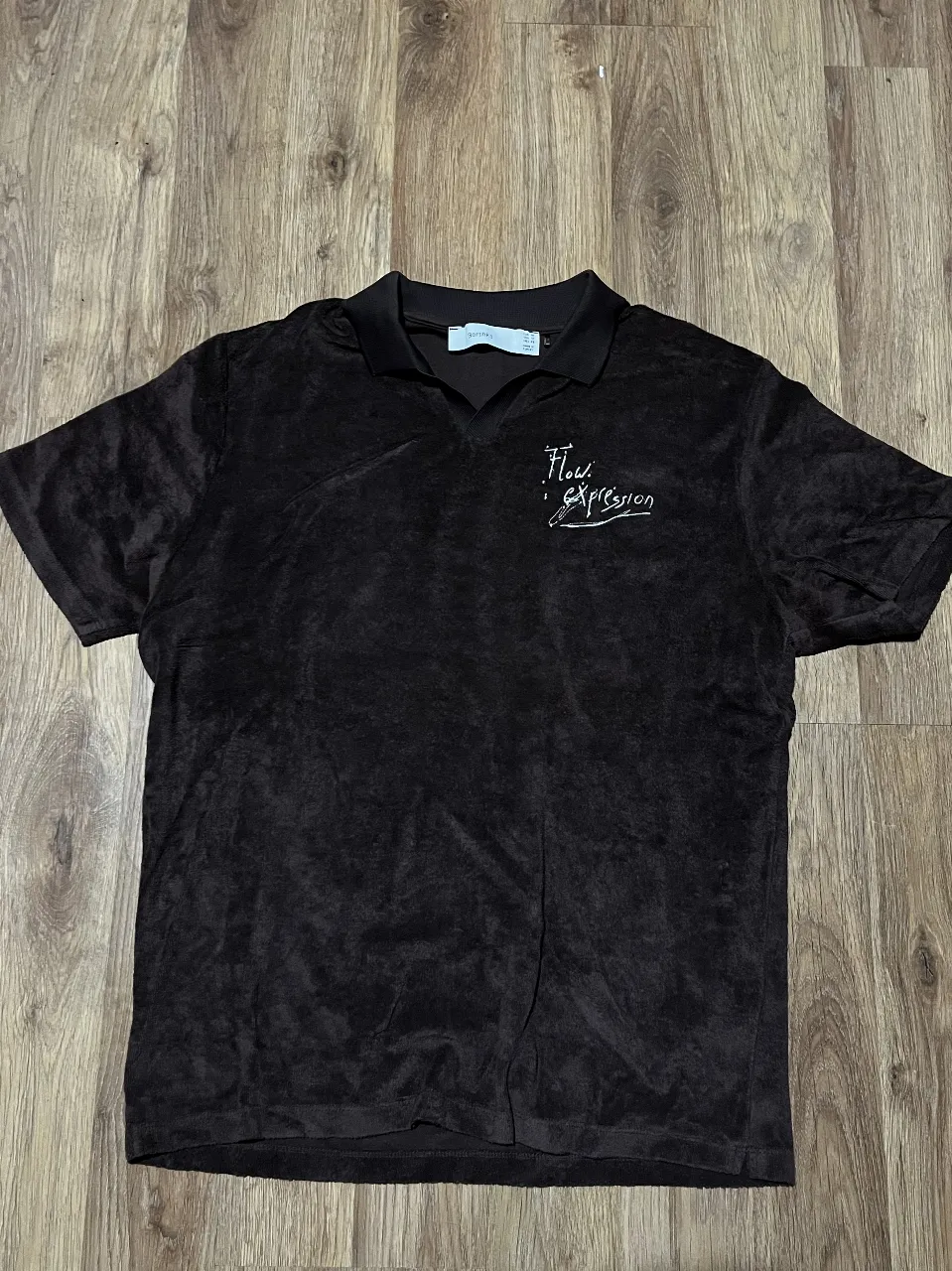 Remera tipo polo de manga corta, confeccionada en un suave tejido tipo toalla. Presenta un diseño liso en color marrón oscuro, con un sutil bordado en el frente que dice "Flow Expression". Ideal para un look casual y cómodo.