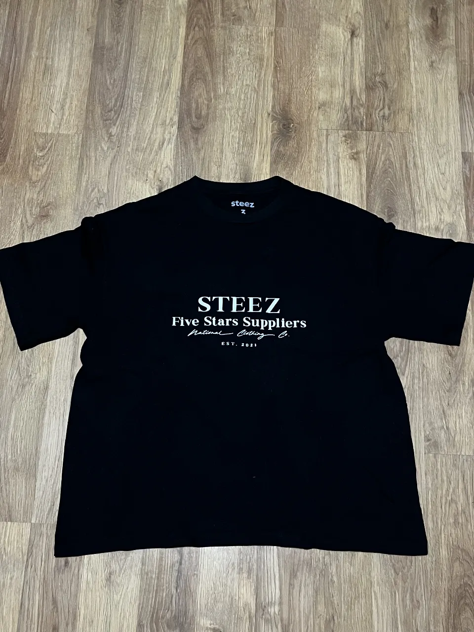 Remera negra de algodón con estampa "STEEZ Five Stars Suppliers National Clothing Co. EST. 2021" en blanco. Cuello redondo y mangas cortas. Perfecta para un look casual y urbano.