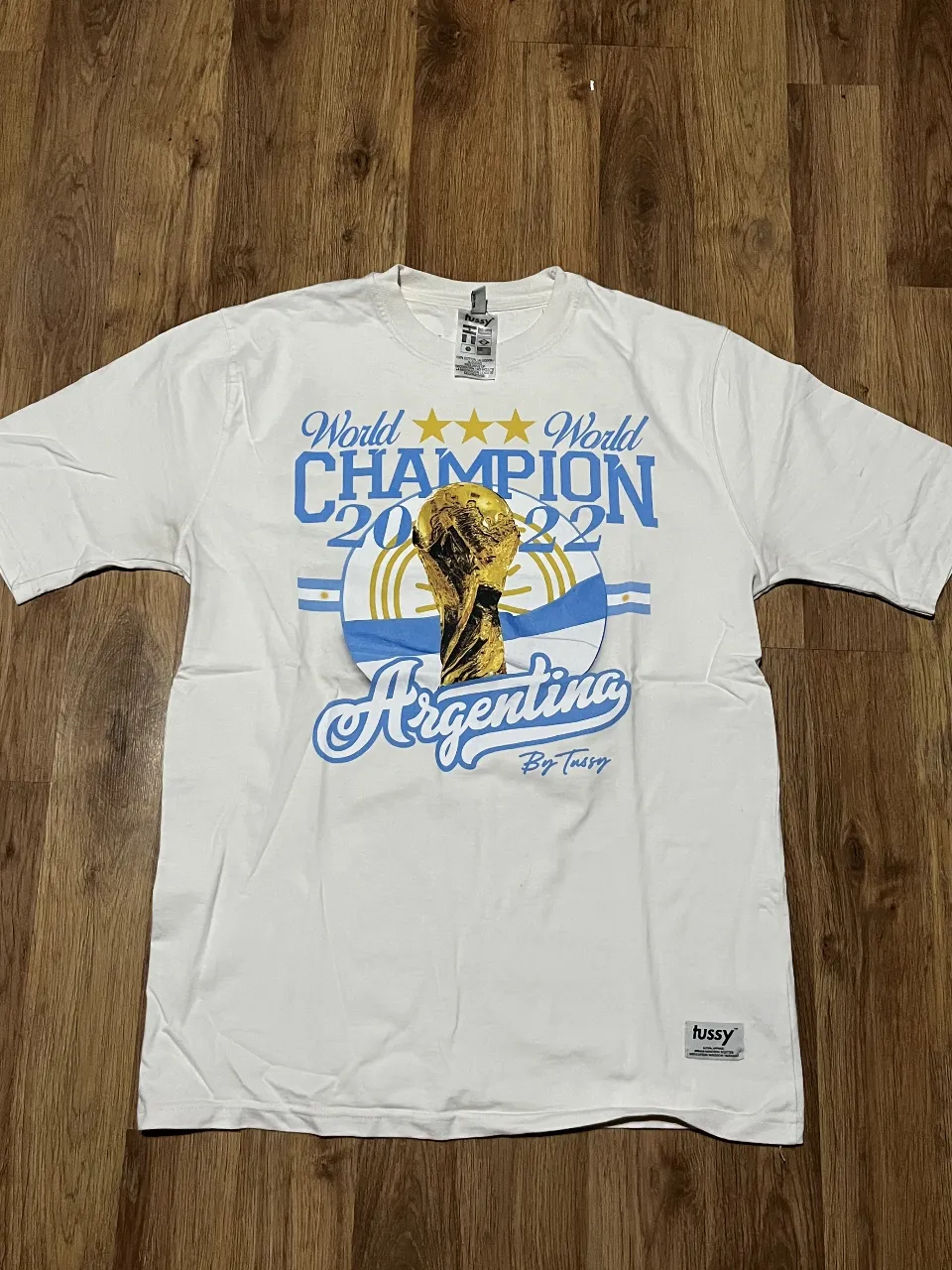 Remera blanca Tussy de algodón con estampa del Mundial Qatar 2022, celebrando a Argentina como campeón. Presenta el trofeo de la Copa del Mundo, estrellas doradas y el nombre "Argentina" en letras celestes. Ideal para los fanáticos del fútbol y para lucir con orgullo los colores de la selección.