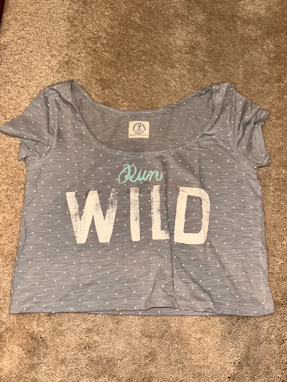 Remera de mujer American Eagle, gris con lunares y estampado "Run Wild". Corte crop, ideal para un look casual y cómodo.