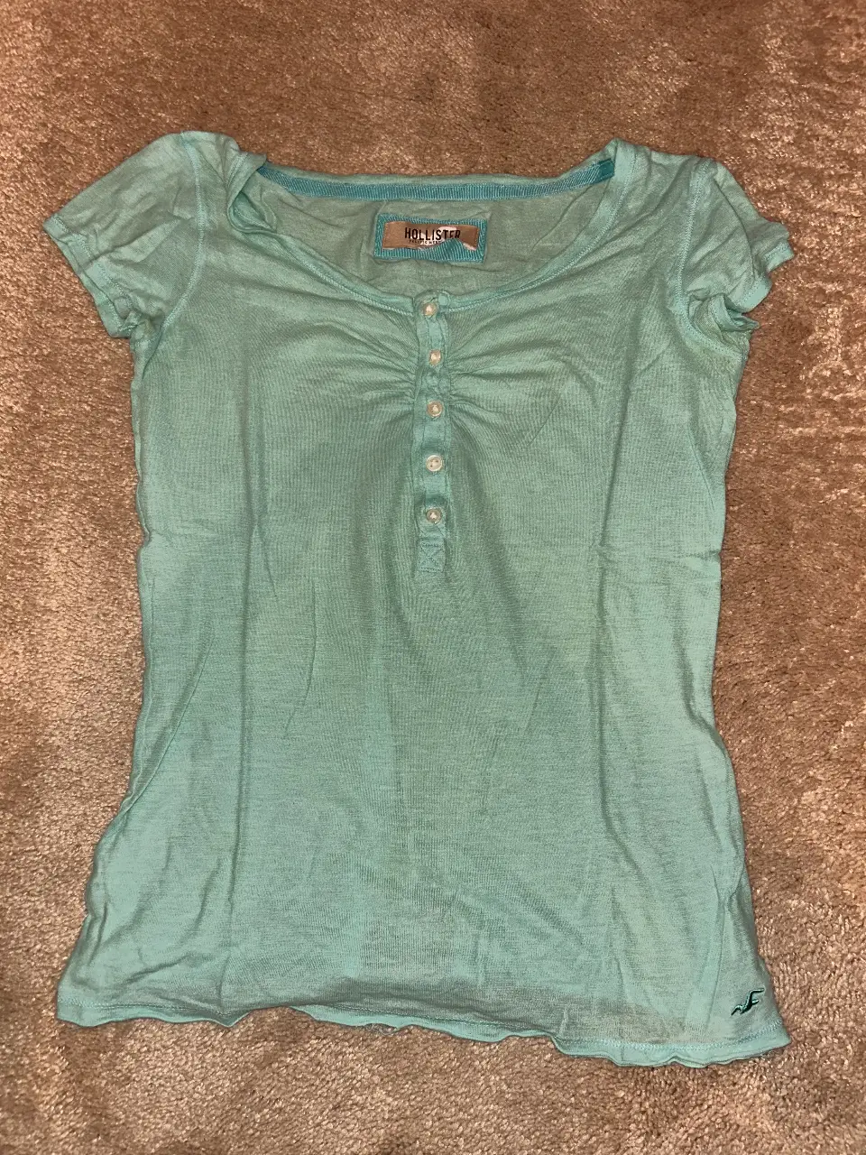 Remera de mujer Hollister, manga corta, color celeste, con botones y detalle fruncido en el frente. Ideal para un look casual y fresco.