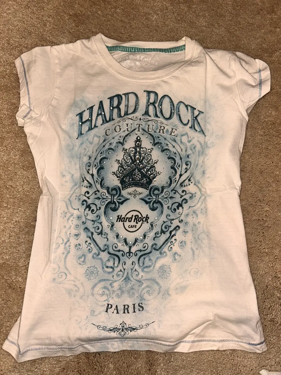 Remera blanca de Hard Rock Cafe Paris para mujer. Con estampado frontal de Hard Rock Couture y un diseño con detalles en azul. Cuello redondo y mangas cortas. Ideal para un look casual y con estilo.