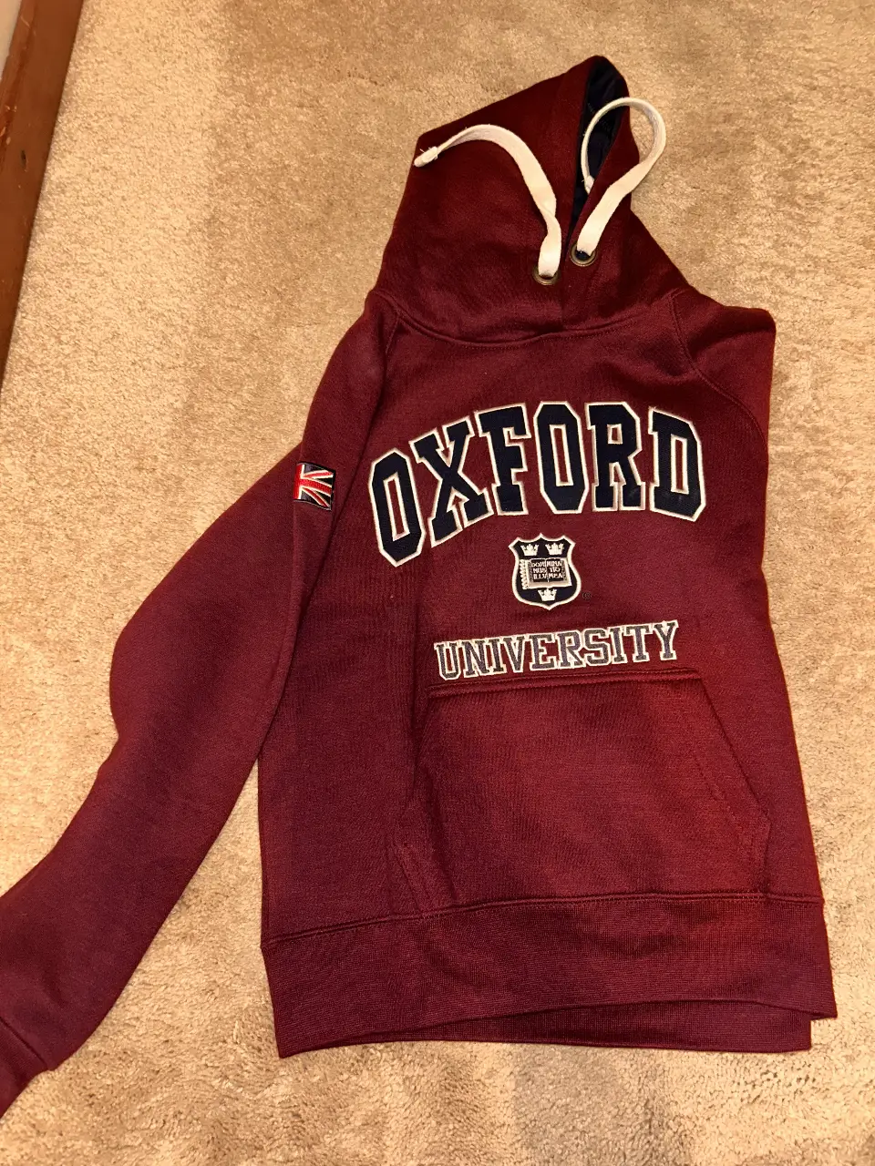 Buzo canguro Oxford University, color bordó, con capucha y bolsillo frontal. Con logo de la universidad estampado en el frente y detalle de bandera en la manga. Ideal para un look casual y universitario.