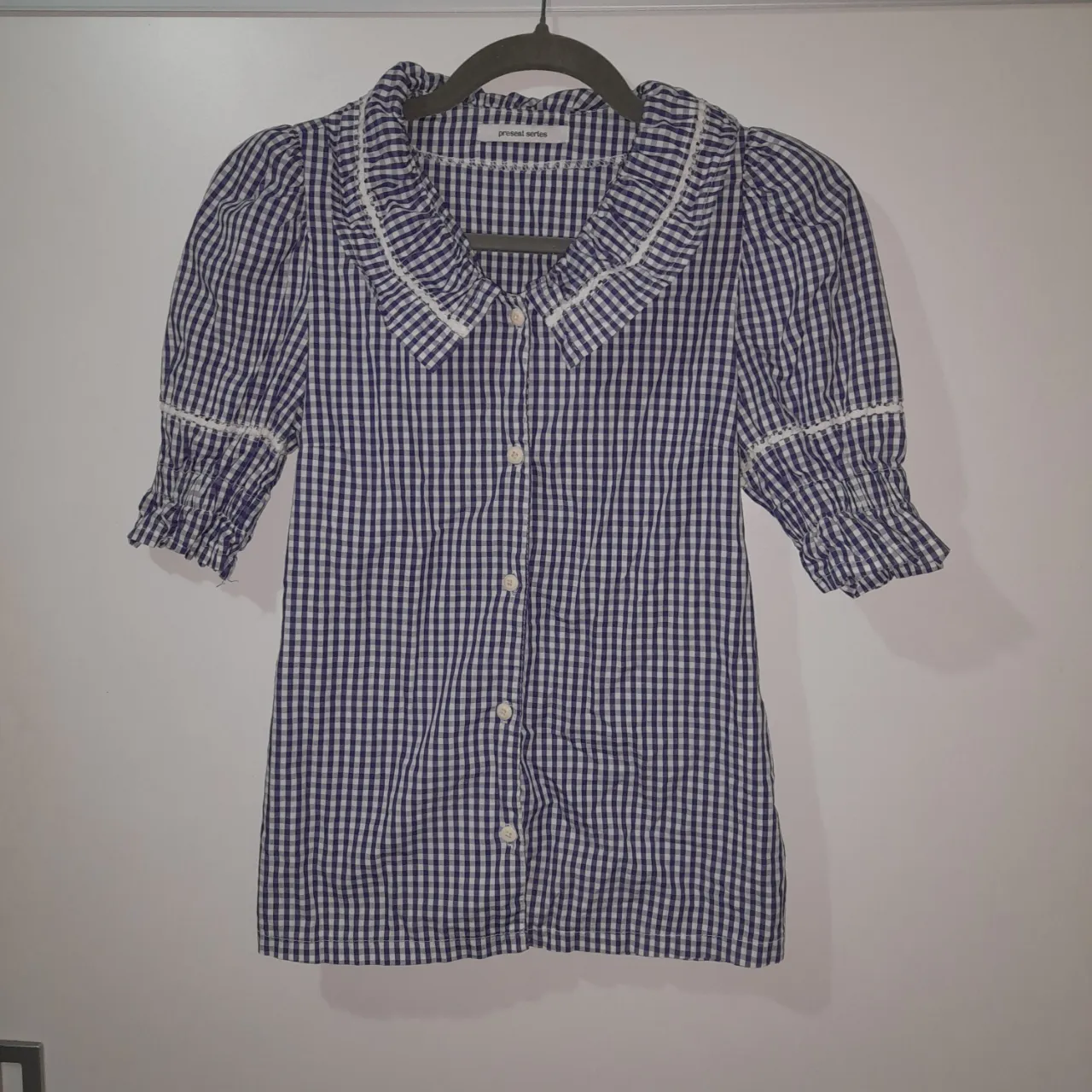 Camisa a cuadros en azul y blanco. Cuello volado con detalle de puntilla blanca. Mangas cortas abullonadas con elástico y detalle de puntilla blanca. Botones al frente.