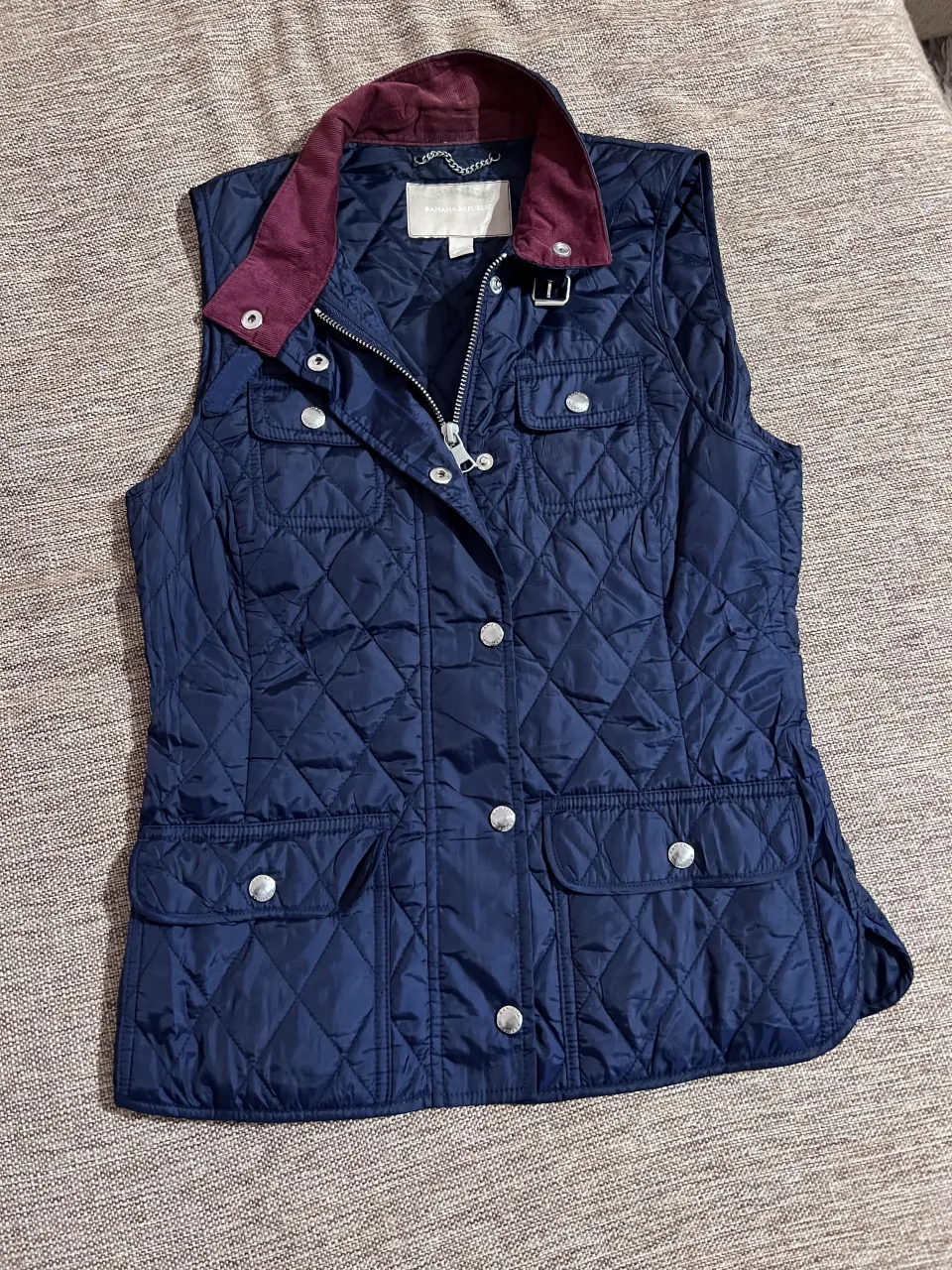 Chaleco acolchado azul marino con cuello de pana bordó. Posee cierre y botones a presión al frente, dos bolsillos superiores con botón y dos bolsillos inferiores con solapa y botón. Detalle de ajuste con botón en la cintura en la parte trasera.