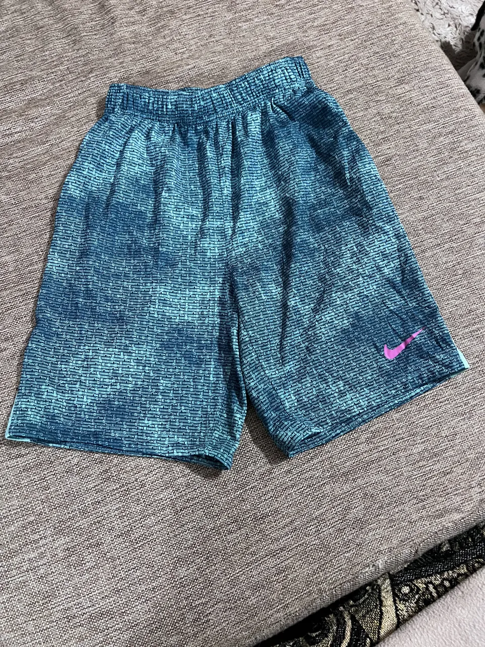 Shorts deportivos Nike con estampado de logo repetido en tonos de azul y verde. Cintura elástica con cordón interno. Logo de Nike en rosa en la pierna derecha.