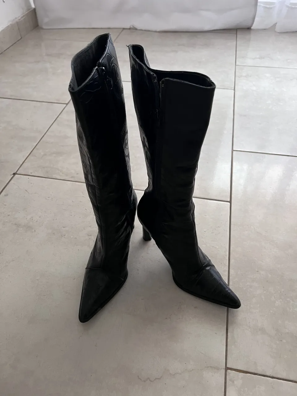 Botas negras de caña alta con taco fino y punta fina. Tienen cierre lateral y un diseño grabado en toda la superficie.