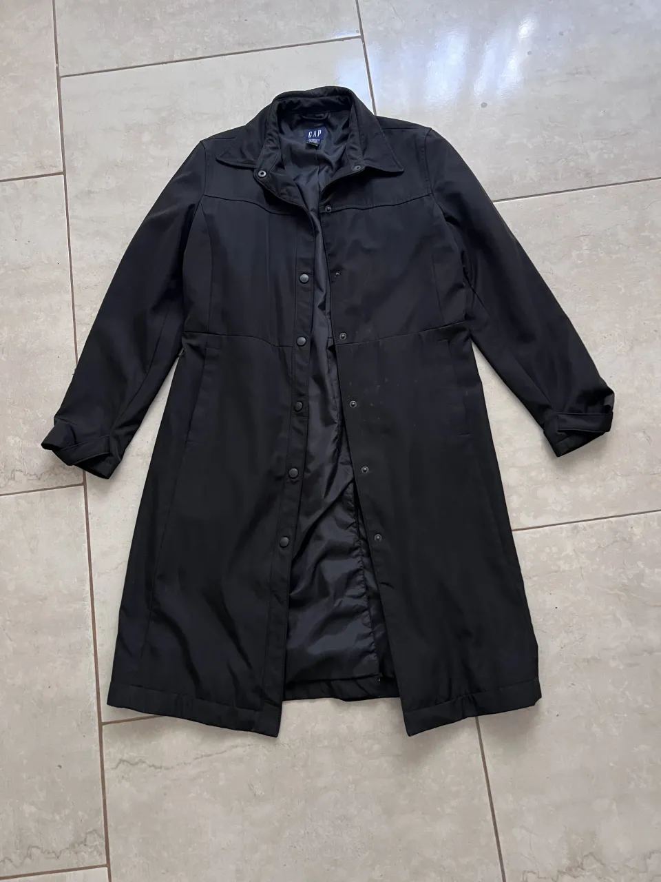 Campera de gabardina negra, larga, con botones al frente y cuello camisero. Tiene bolsillos laterales.