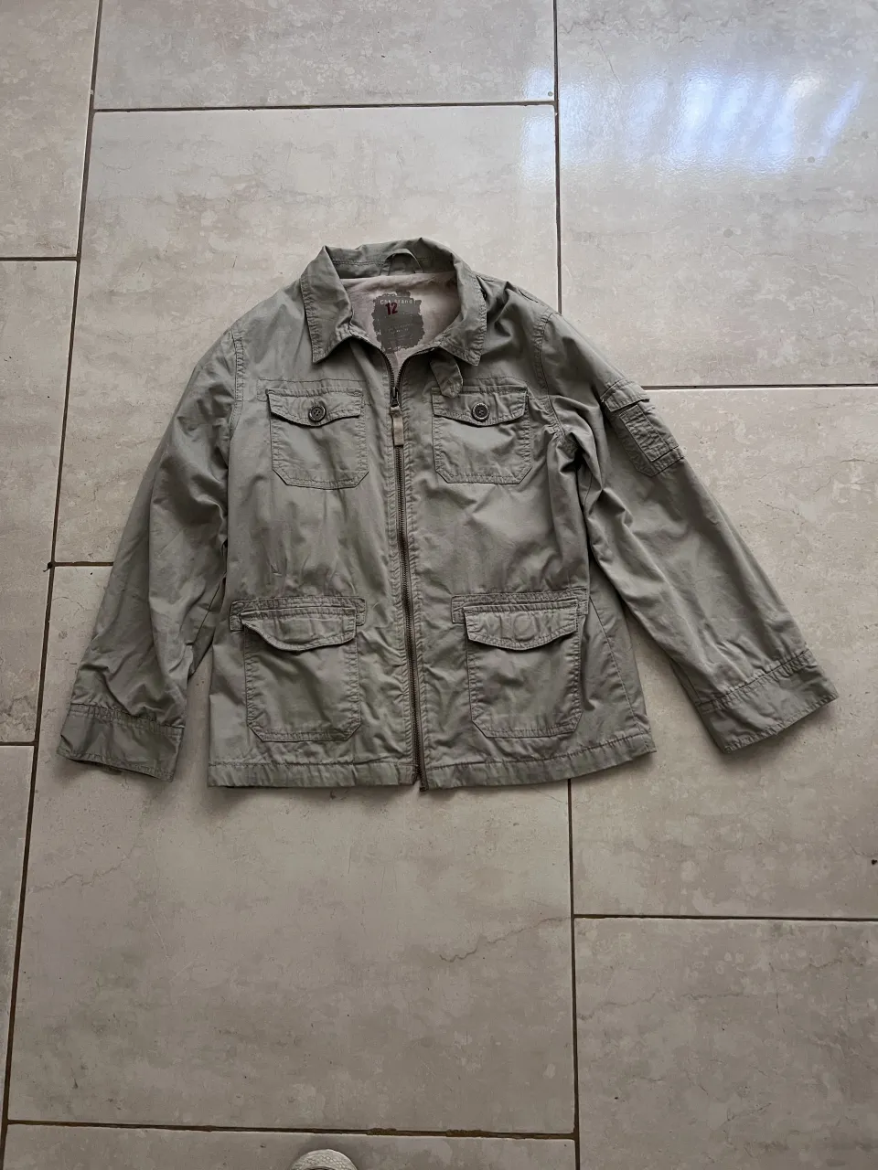 Chaqueta de color verde militar con cuello camisero, cierre completo y cuatro bolsillos frontales con tapa y botón. Posee un bolsillo adicional con tapa en la manga izquierda.