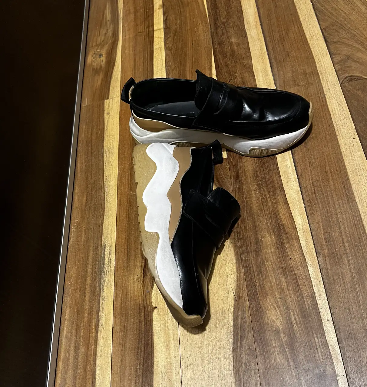 Zapatillas tipo mocasín de cuero negro con suela gruesa de goma blanca y detalles en beige. Un calzado moderno y versátil, ideal para un look casual con un toque de sofisticación.