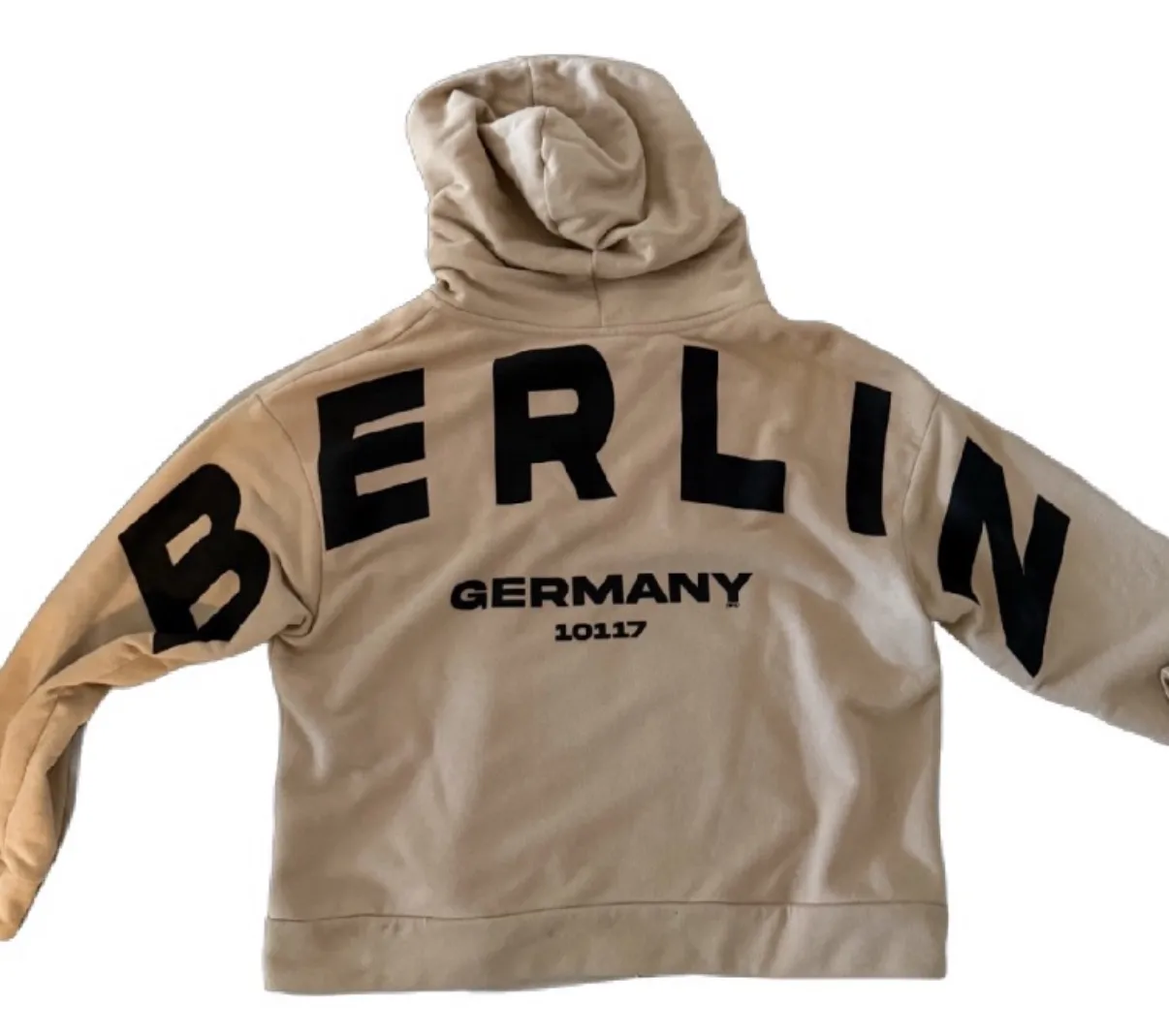 Buzo con capucha y cierre completo, color beige, con estampa grande de "BERLIN GERMANY 10117" en la espalda. Tiene detalles de franjas negras en las mangas. Ideal para un look urbano y casual.