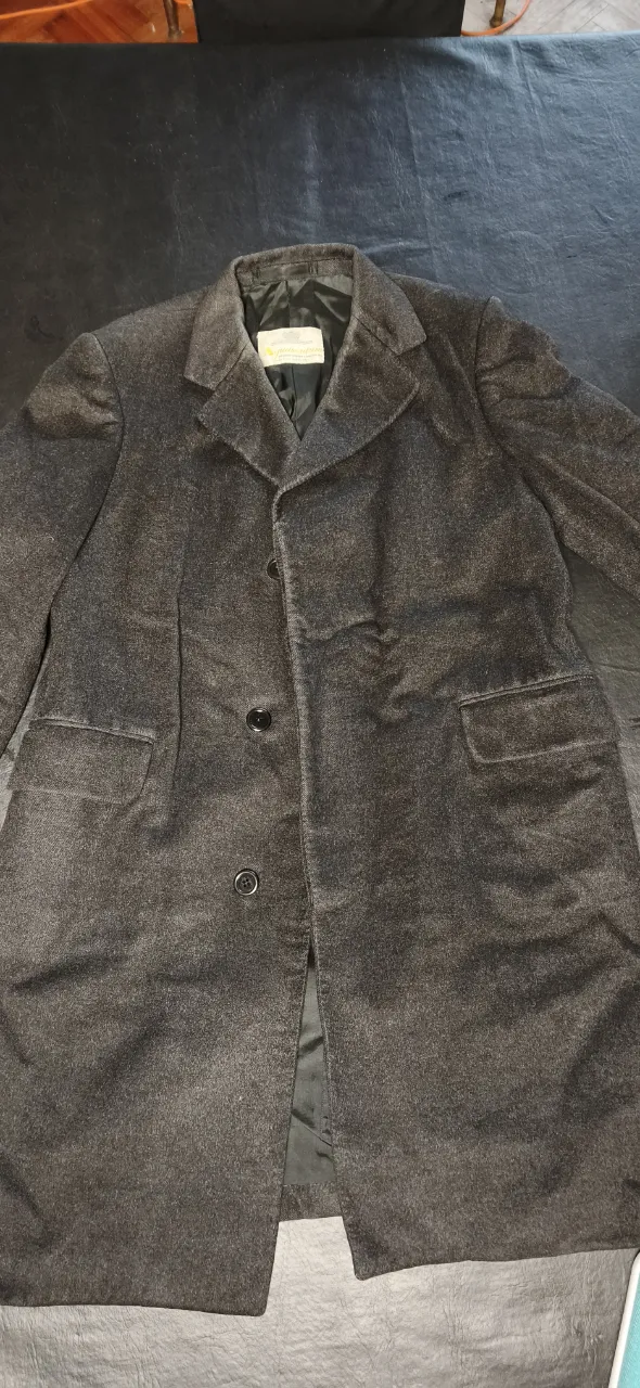 Abrigo largo de cashmere color negro. Posee cuello solapa, tres botones al frente y dos bolsillos con tapa. El interior es forrado en tela lisa negra. Pequeños detalles de uso, una pequeña parte inferior está descosida, y una pequeña rotura en la axila izquierda, por dentro.