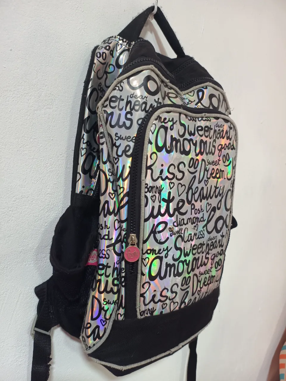 Mochila súper canchera con un diseño holográfico lleno de inscripciones como 'sweet heart', 'amorous', 'kiss', 'beauty' y 'cute'. Tiene detalles en negro y un charm rosa en el cierre. Ideal para darle un toque divertido y original a tu look.
