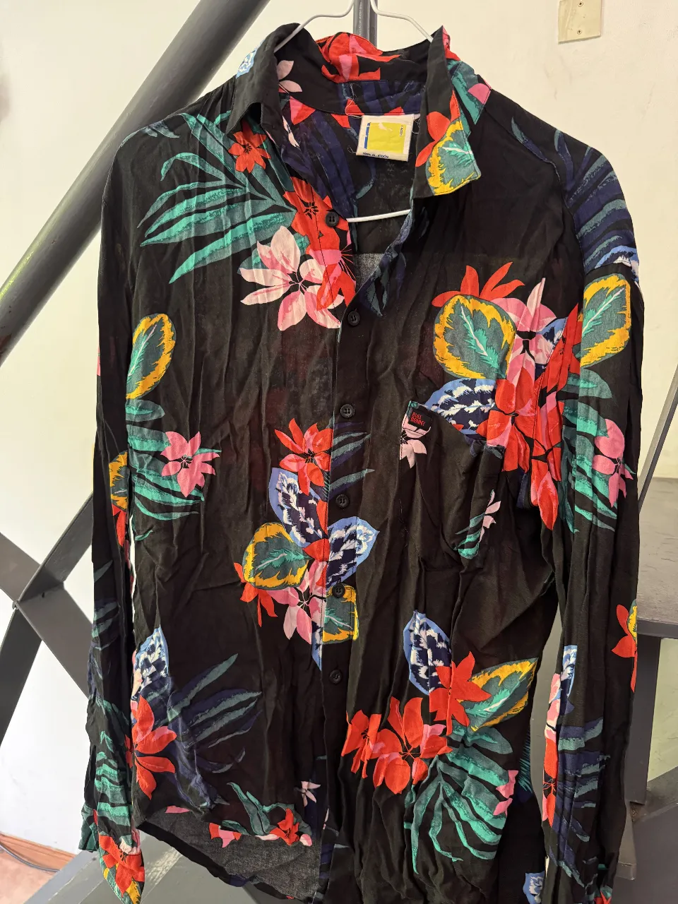 Camisa de manga larga con estampa tropical vibrante. Fondo negro con flores y hojas en colores llamativos como rojo, rosa, amarillo, verde y azul. 