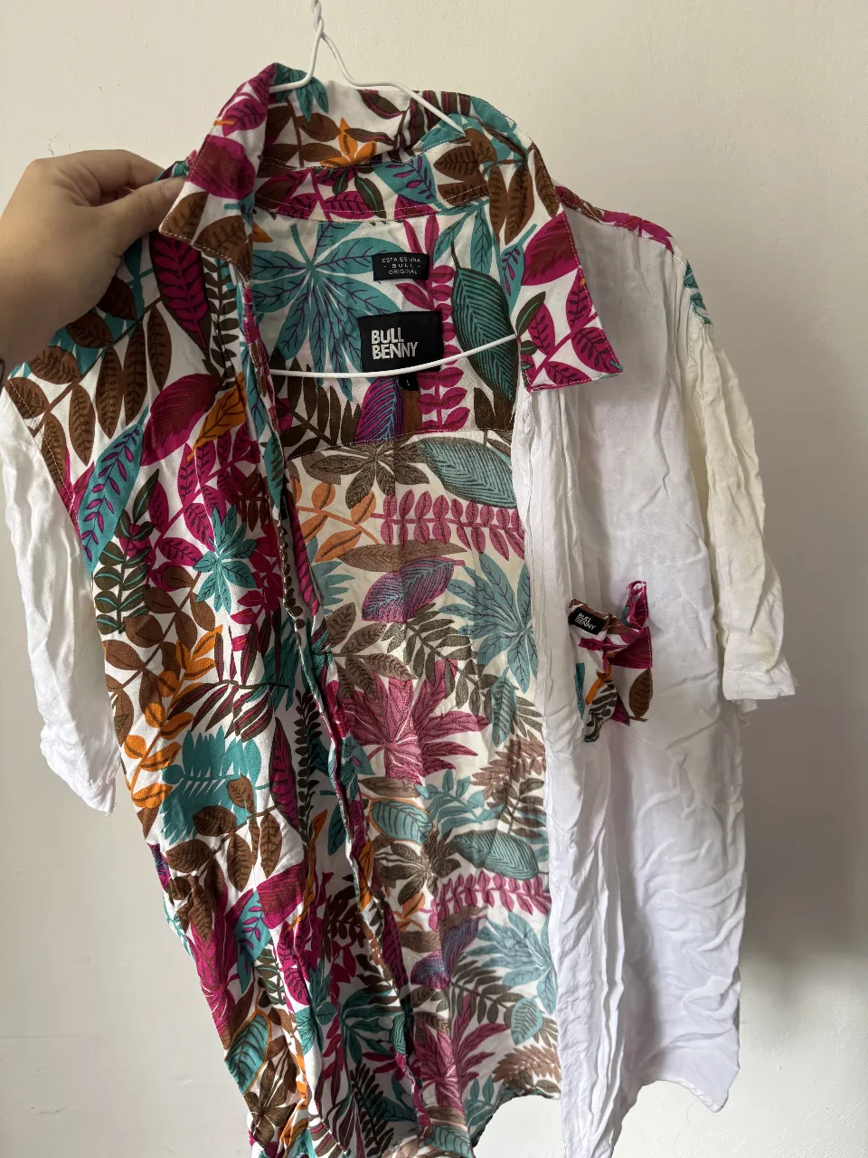 Camisa de manga corta con un diseño vibrante y tropical. Presenta un estampado de hojas y flores en colores vivos como fucsia, turquesa, naranja y marrón sobre un fondo blanco en la parte delantera y una manga. 