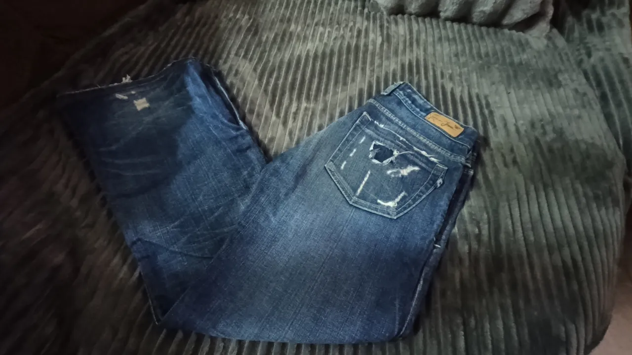 Jeans de denim azul con roturas y desgastes. Presenta detalles de costuras visibles y parches en los bolsillos traseros.