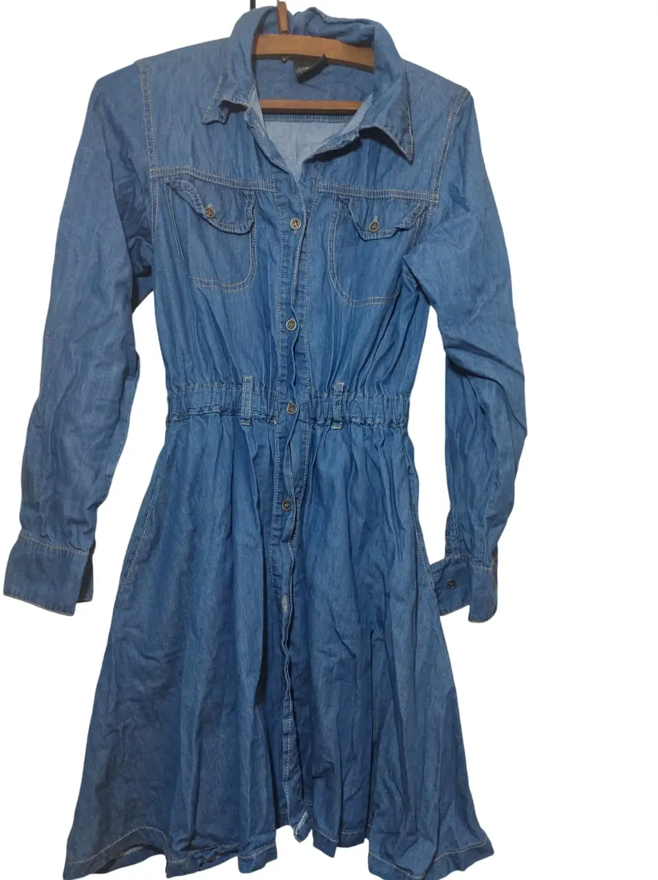 Vestido de jean azul, con mangas largas y cintura elastizada. Posee botones frontales y dos bolsillos en el pecho. Ideal para un look casual y cómodo.