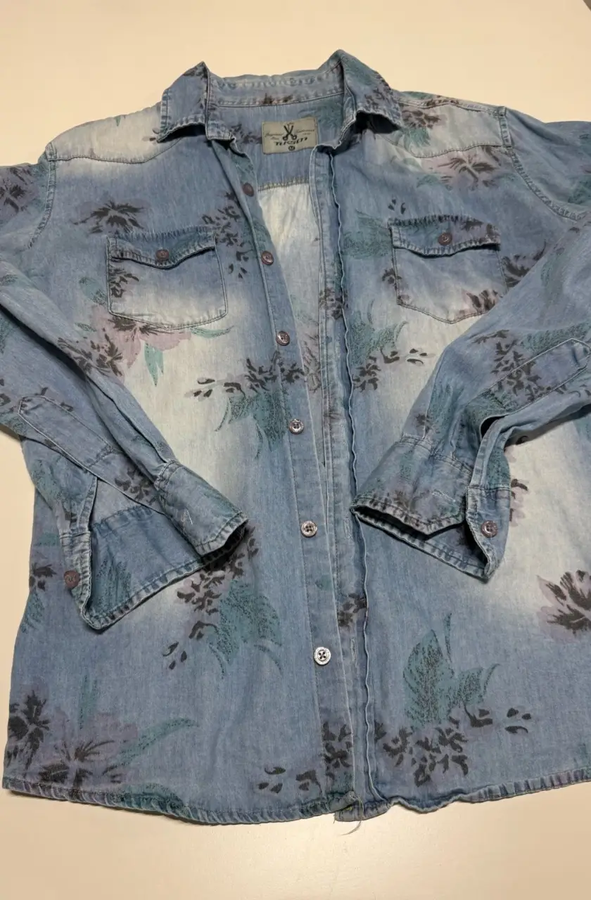Camisa de jean celeste con estampado floral en tonos grises y lilas. Corte clásico, con dos bolsillos delanteros y botones. Ideal para un look casual y canchero. Largo: 68. Sisa: 50 cm (contorno: 100 cm)