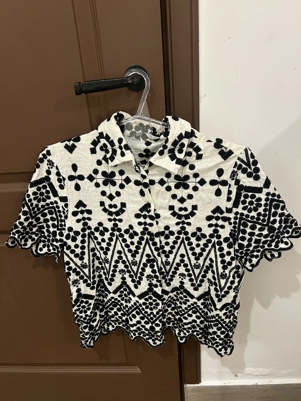 Camisa blanca con bordados negros y mangas cortas. Posee un diseño de zigzag y detalles florales en toda la prenda, con un corte moderno y fresco. Ideal para un look casual y chic.