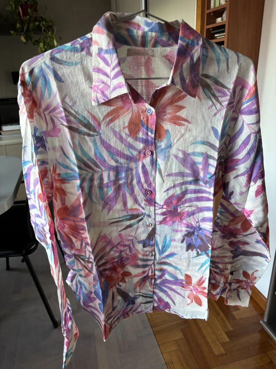 Camisa de manga larga con estampado floral y de hojas en tonos vibrantes de rosa, violeta, azul y naranja sobre fondo blanco. Posee cuello clásico y botones al frente.