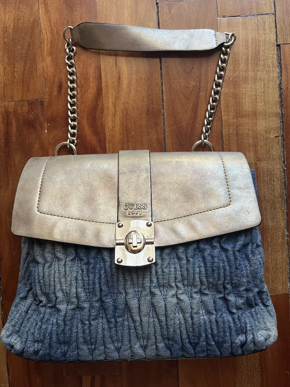 Cartera Guess 1981, con diseño de denim fruncido y solapa dorada. Detalles de cadena y cierre con logo. Ideal para un look casual y elegante.