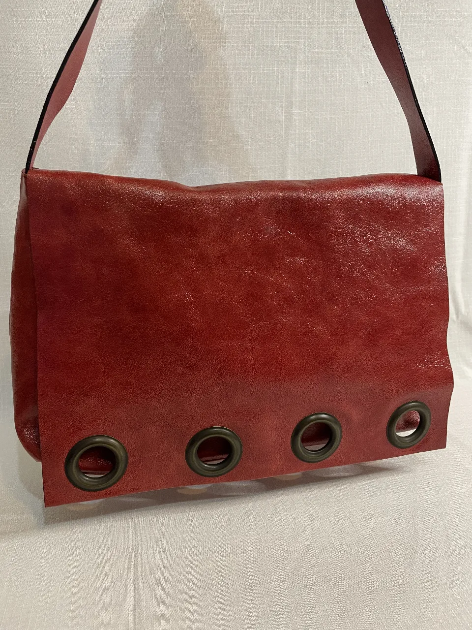 Cartera bandolera de cuero rojo con detalles de ojales metálicos. Diseño moderno y funcional, ideal para llevar tus esenciales con estilo. Interior forrado en tela violeta.