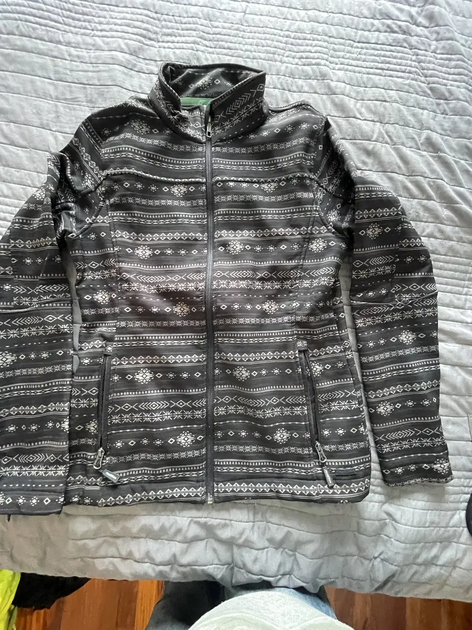 Campera deportiva con cierre y estampado geométrico en tonos grises y blancos. Ideal para actividades al aire libre o uso casual. Posee bolsillos laterales con cierre y cuello alto.