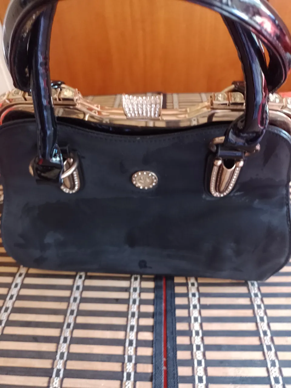 Cartera negra con manijas cortas y detalles metálicos dorados y plateados. Posee un aplique de strass en la parte superior y un broche.