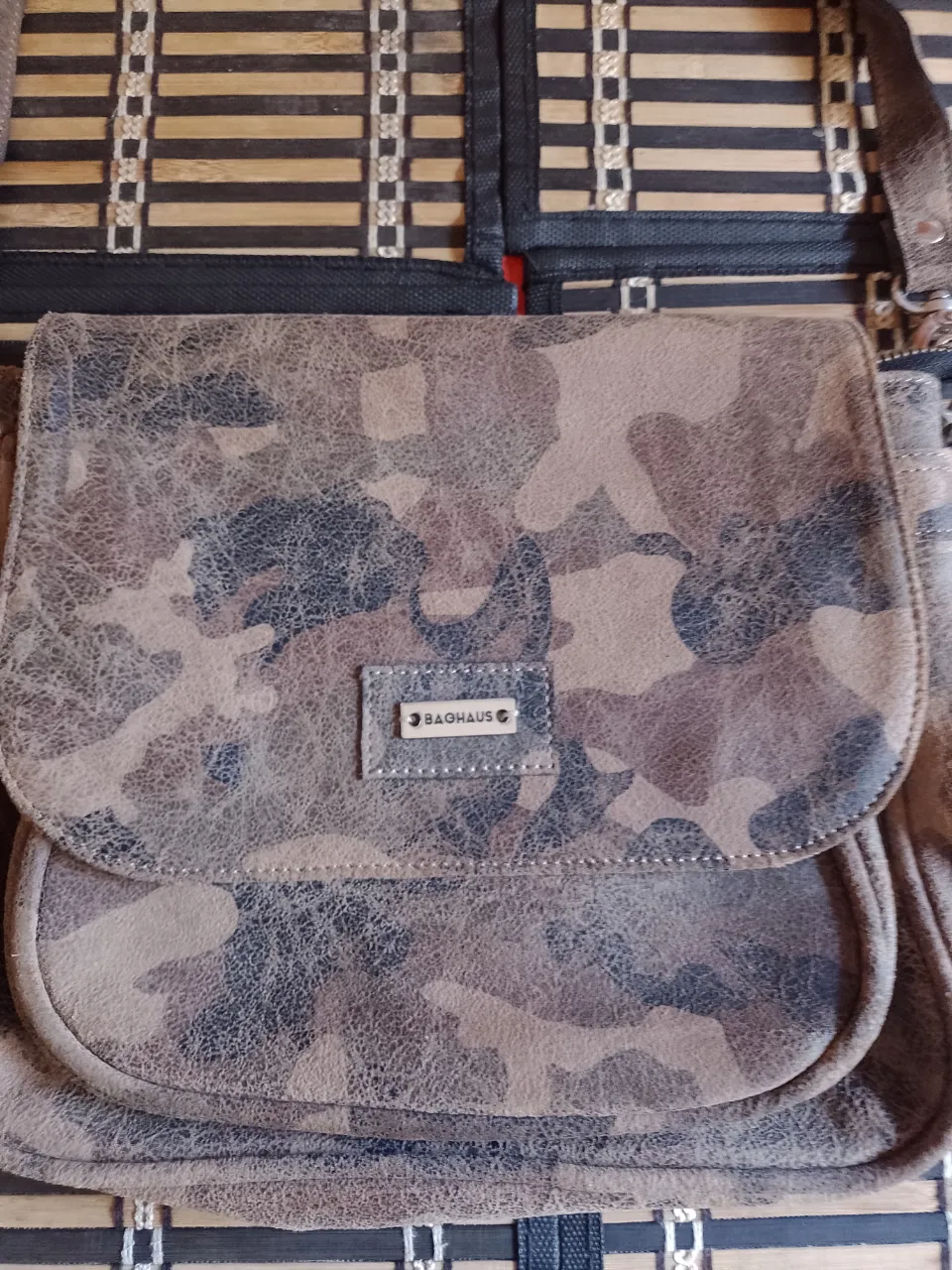 Cartera con estampa camuflada en tonos marrones y grises. Posee una solapa con cierre de botón a presión y un bolsillo frontal. La correa es ajustable.