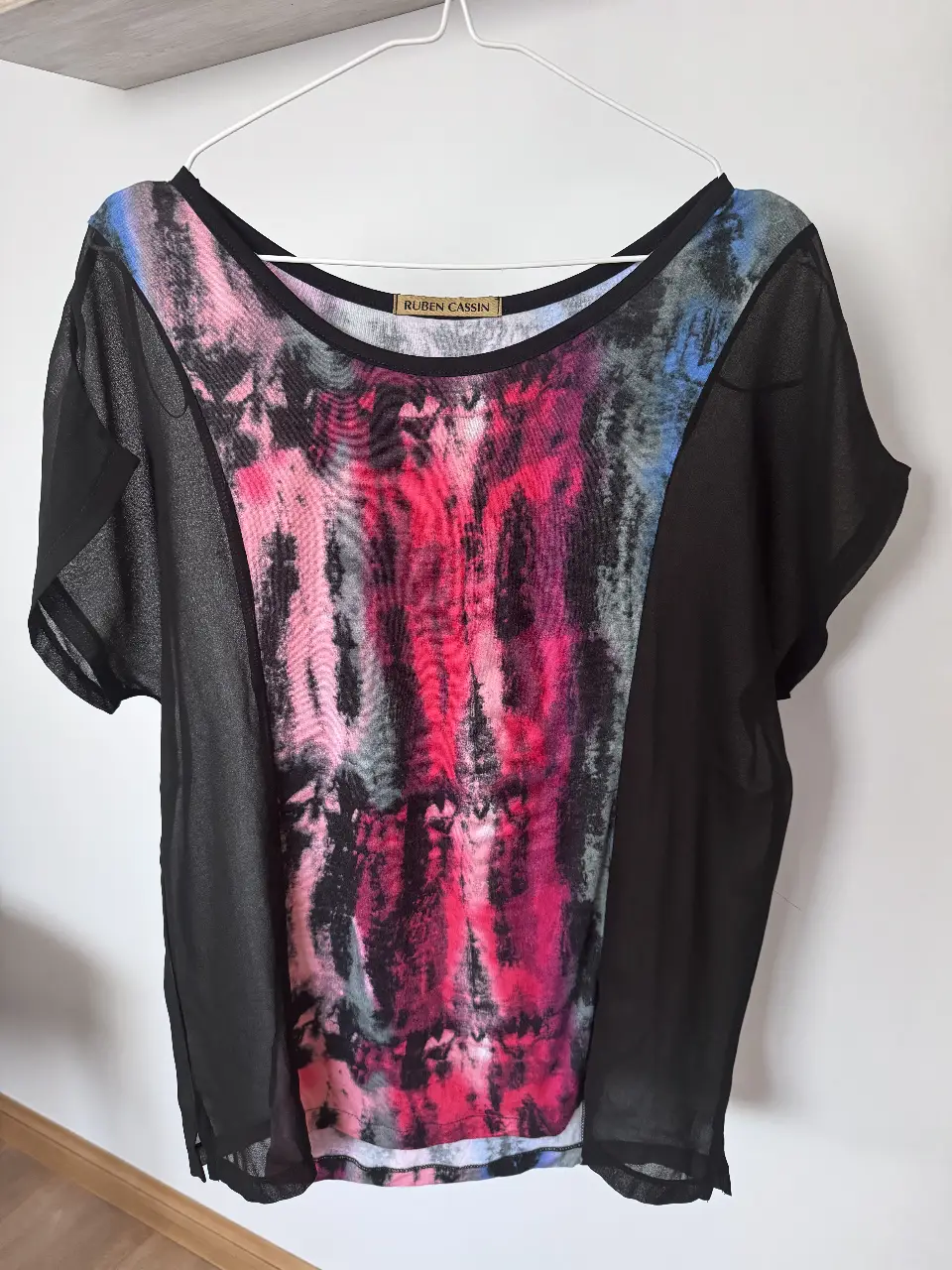 Blusa de mujer con estampado abstracto en tonos rosa, rojo y azul grisáceo. Mangas cortas y paneles laterales negros transparentes. Ideal para un look casual y moderno.