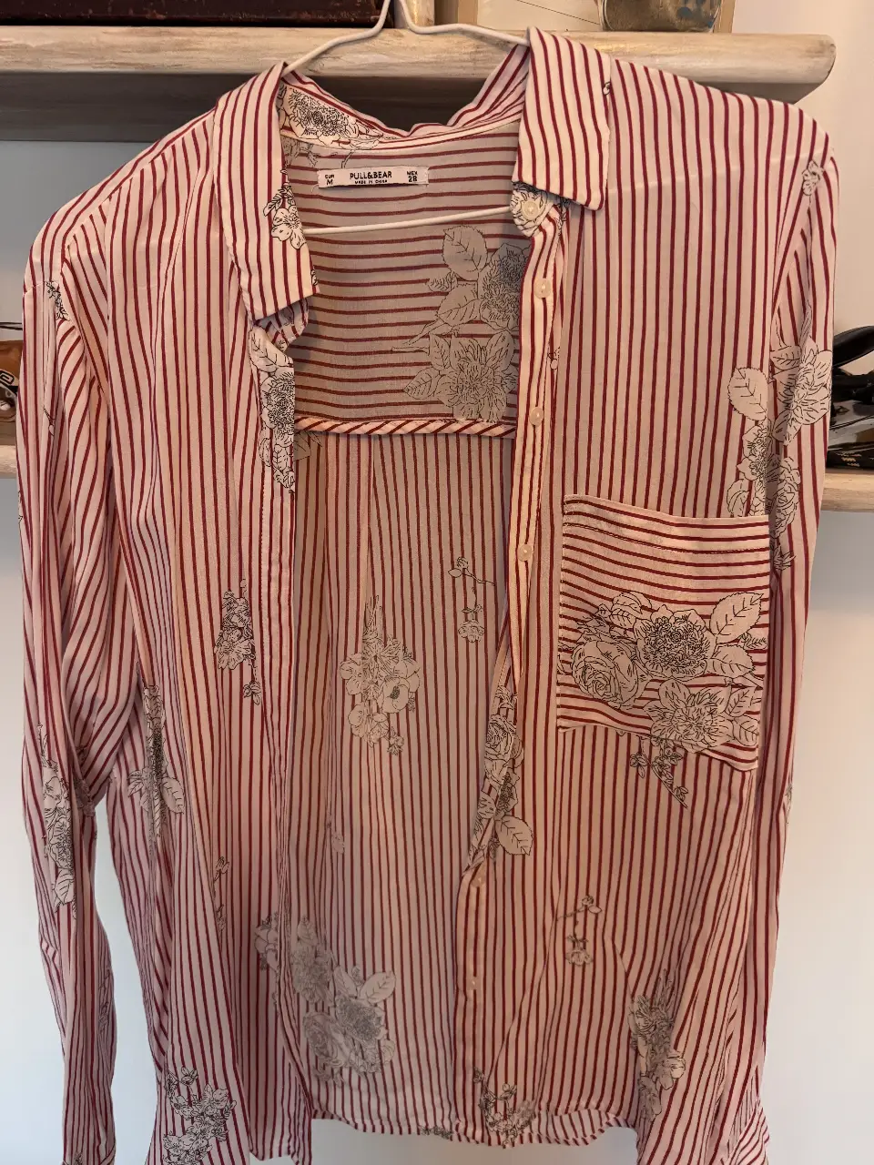 Camisa a rayas rojas y blancas con estampado floral. De manga larga, con bolsillo en el pecho y cuello clásico. Ideal para un look casual y fresco.