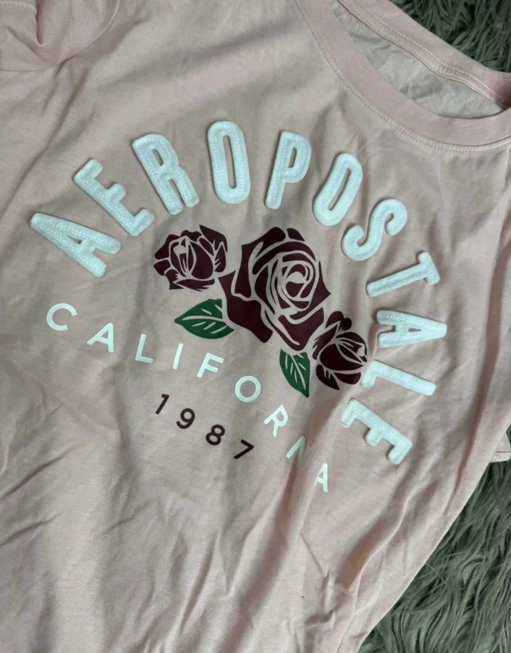 Remera de manga corta Aeropostale para mujer, de color rosa claro, con estampado frontal de la marca Aeropostale, con diseño de rosas y letras en relieve. Ideal para un look casual y cómodo.