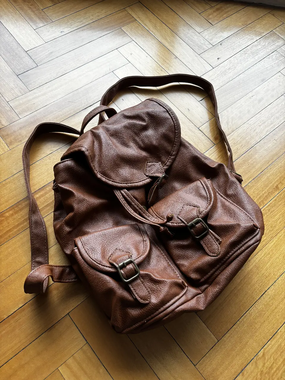 Mochila marrón de cuero sintético con dos bolsillos frontales con hebillas y solapa superior. Ideal para uso diario y un estilo casual.