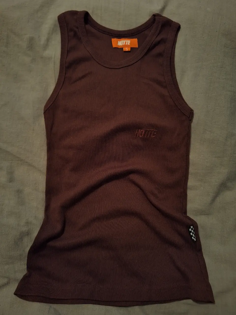 Musculosa color marrón, de tejido acanalado. Tiene el logo de la marca "NOTTE" bordado en el pecho y una etiqueta en el cuello. Ideal para un look casual y cómodo.