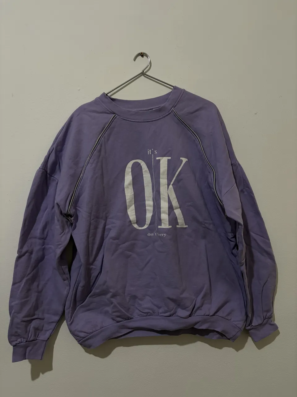 Buzo oversize color lila con estampa "it's OK don't worry" en blanco. Tiene detalles de cierre en los hombros y puños elastizados.