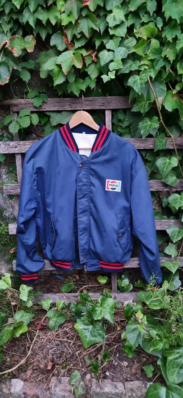 Campera bomber azul marino de Pepsi, con logo en el pecho y estampa grande en la espalda. Cuello y puños con franjas rojas y blancas. Cierre metálico YKK y botones a presión.