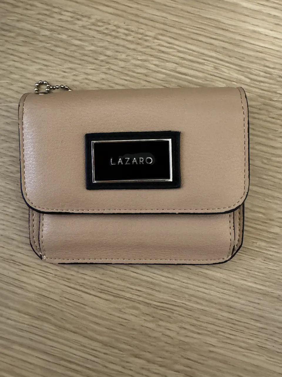 Billetera Lazaro de cuero sintético en tono nude. Diseño compacto con solapa y cierre a presión. Interior con compartimentos para tarjetas y billetes, además de un monedero con cierre. Detalles en negro y plateado.