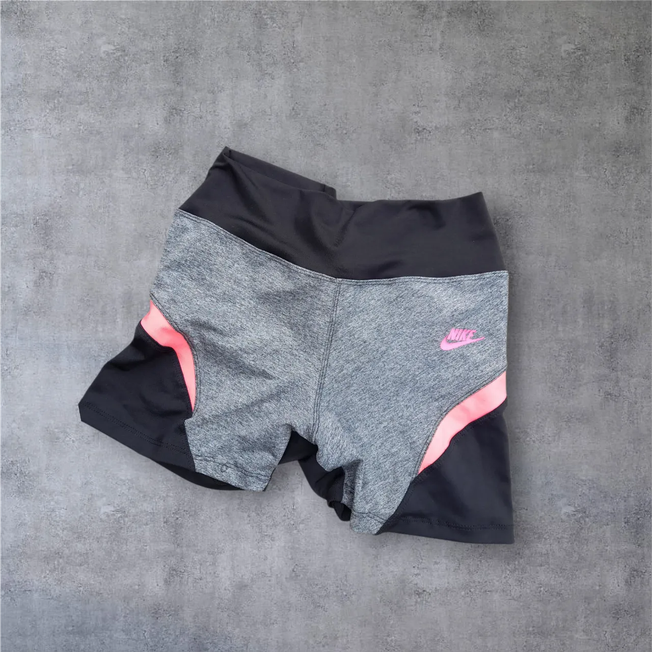 Shorts deportivos Nike, ideales para entrenar o para un look casual y cómodo. Diseño moderno con detalles en rosa vibrante y paneles laterales en negro y gris jaspeado. Cintura alta elastizada para un ajuste perfecto.