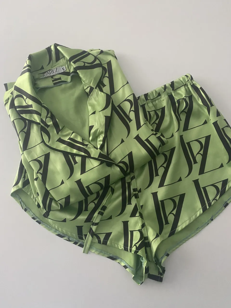 Conjunto de pijama de satén súper suave y fresco, con estampado de logos en negro sobre fondo verde lima. Incluye camisa de manga corta con botones y short con cintura elastizada y cordón. Ideal para estar cómoda y a la moda en casa.