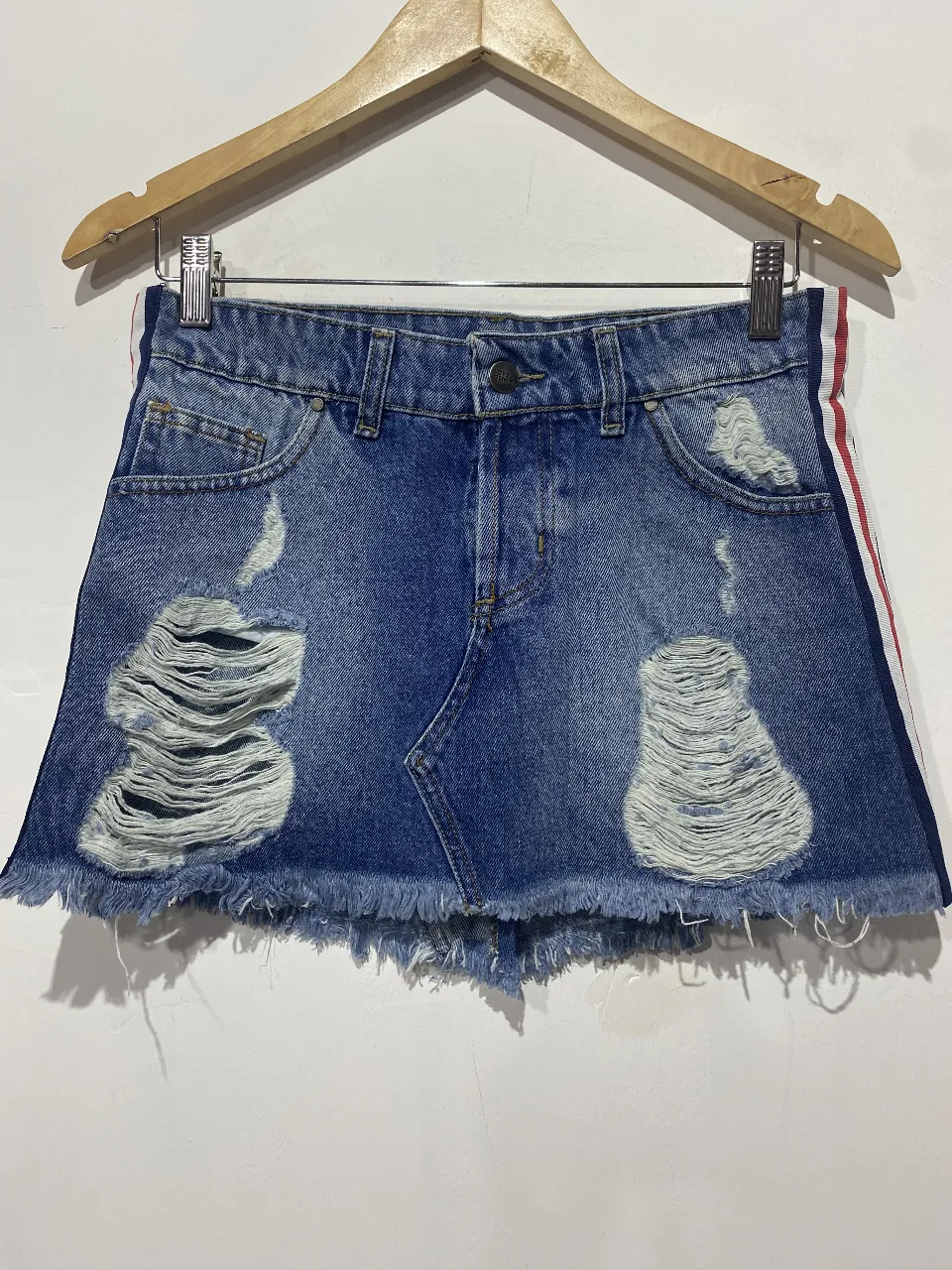 Pollera de jean tiro alto, con roturas desgastadas y detalles de flecos en el ruedo. Posee tiras laterales con los colores de la bandera argentina. Ideal para un look canchero y moderno.