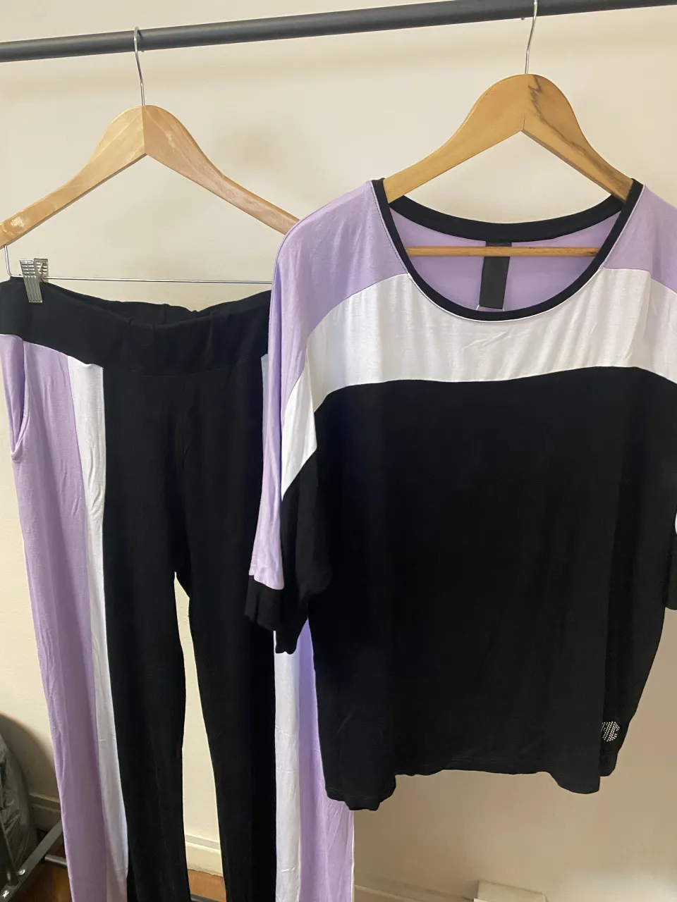 Conjunto deportivo de remera y pantalón de friza. La remera es de mangas 3/4, con bloques de color en negro, blanco y lila. El pantalón es negro con franjas laterales en blanco y lila. Detalle de strass en la remera.