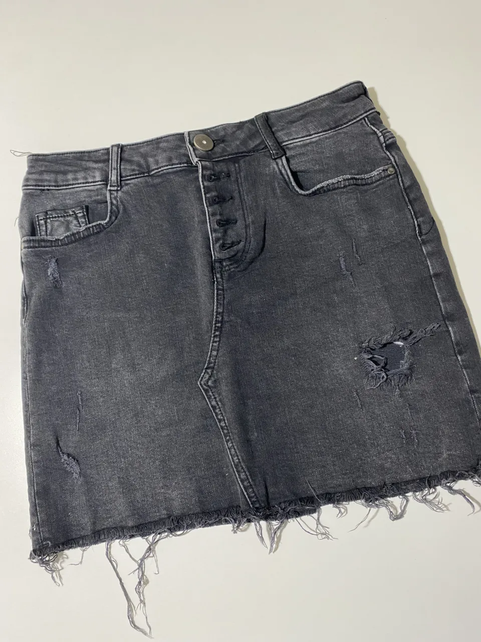 Pollera de jean gris con roturas y detalles deshilachados en el borde inferior. Posee cinco botones en la parte delantera.