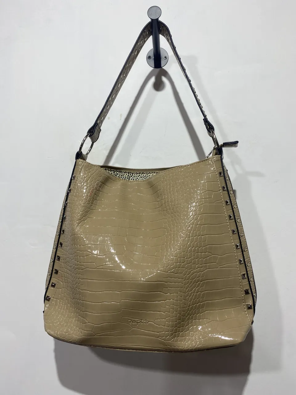 Bolso de cuero sintético con textura de cocodrilo en color beige. Posee tachas metálicas a los costados y un bolsillo exterior con cierre en la parte trasera. El interior está forrado con una tela estampada y cuenta con varios compartimentos.