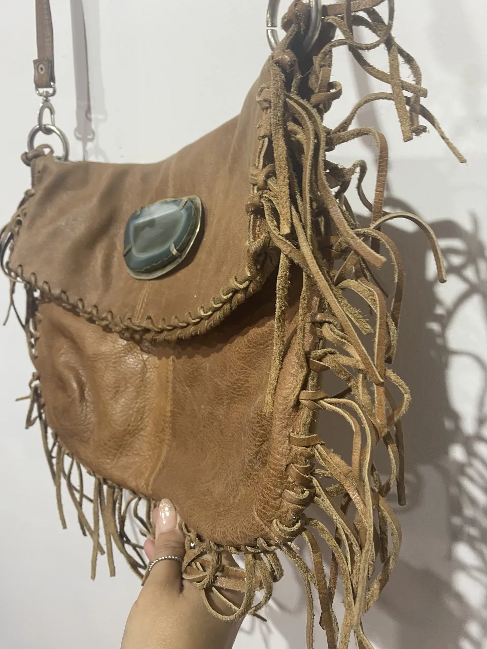 Bolso de cuero marrón con flecos largos y un detalle de piedra natural en la solapa. Tiene un diseño boho y artesanal, ideal para darle un toque único a tu look. Su interior forrado con estampa floral vibrante lo hace aún más especial.