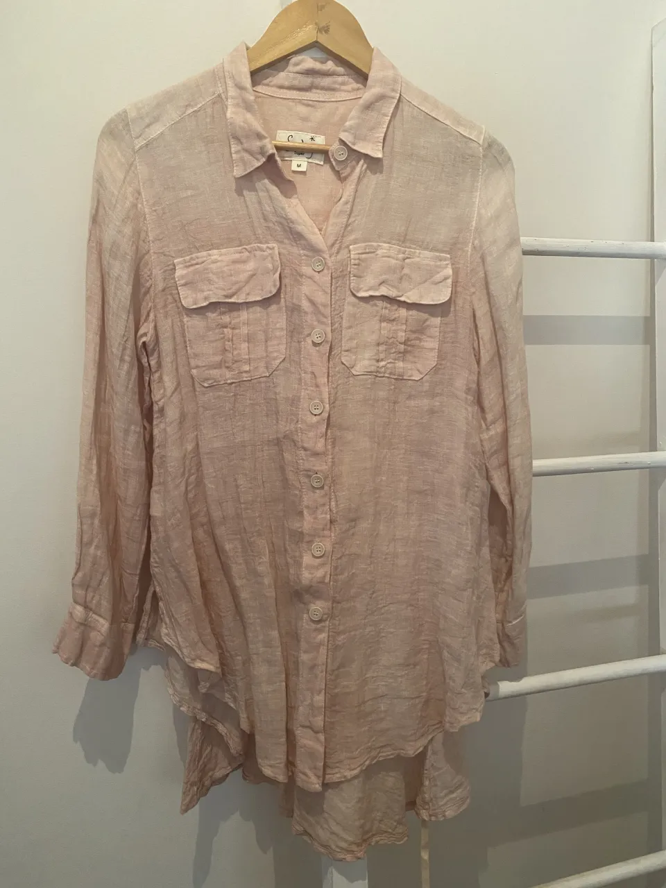 Camisa de lino color rosa pálido, con botones al frente, dos bolsillos en el pecho y mangas largas. Posee un corte holgado y detalles de fruncido en la espalda para ajustar la cintura. Ideal para un look casual y fresco.